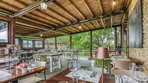 An outdoor dining area with wooden tables and chairs, stone walls, and large windows overlooking greenery.