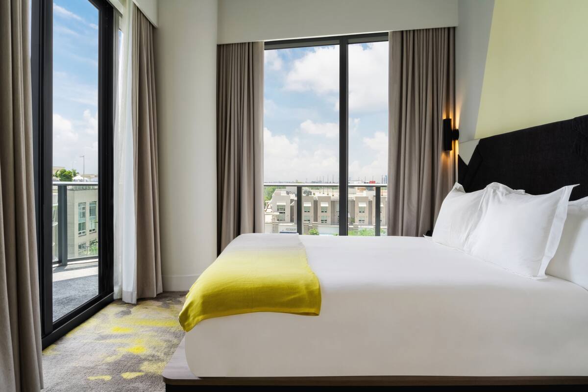 A hotel room with a large bed, a view of the city through a window, and a yellow throw blanket.