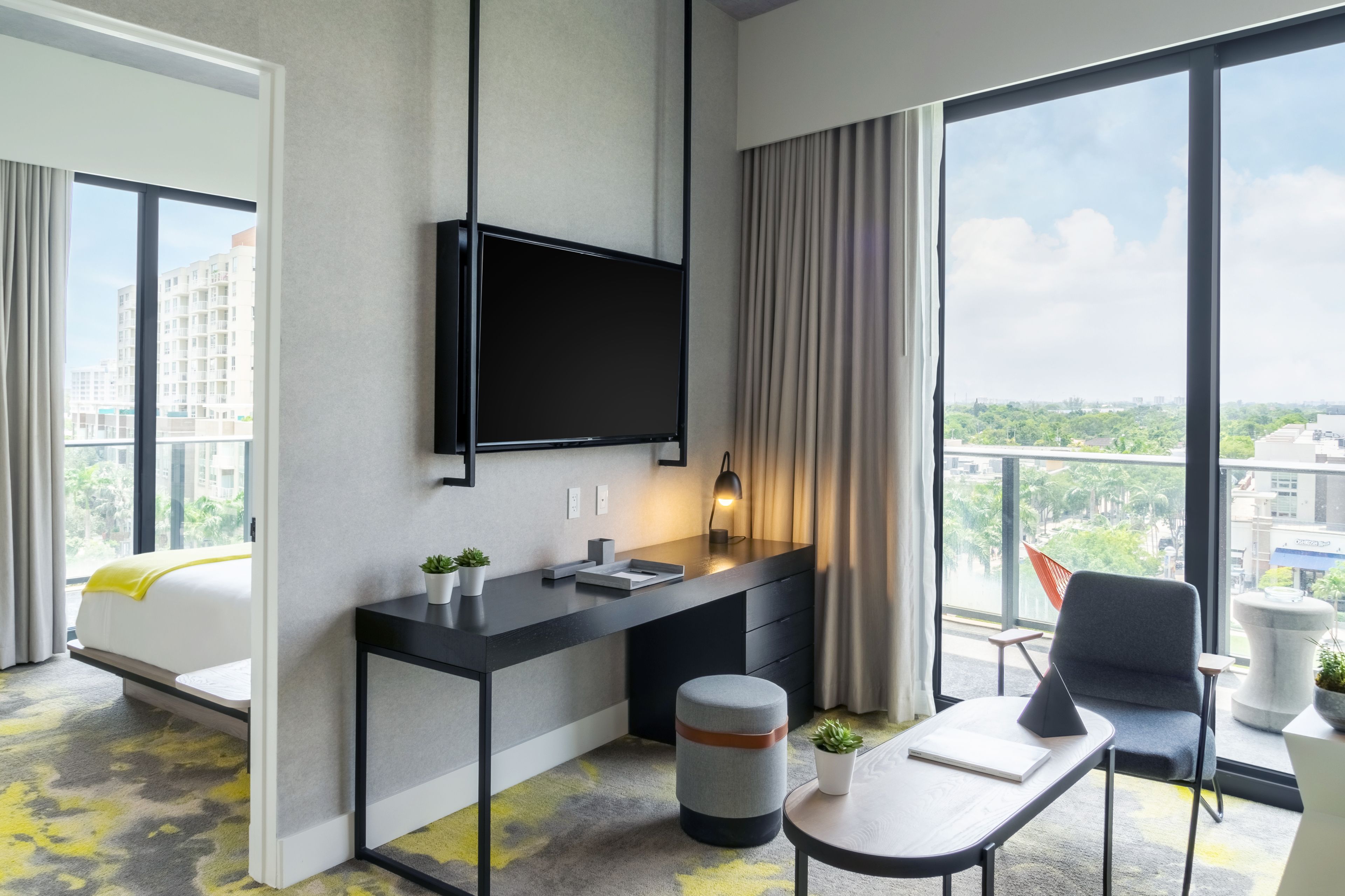 Een moderne hotelkamer met een groot raam, een flatscreen televisie aan de muur, een klein bureau met een lamp, een stoel en een rond poef.