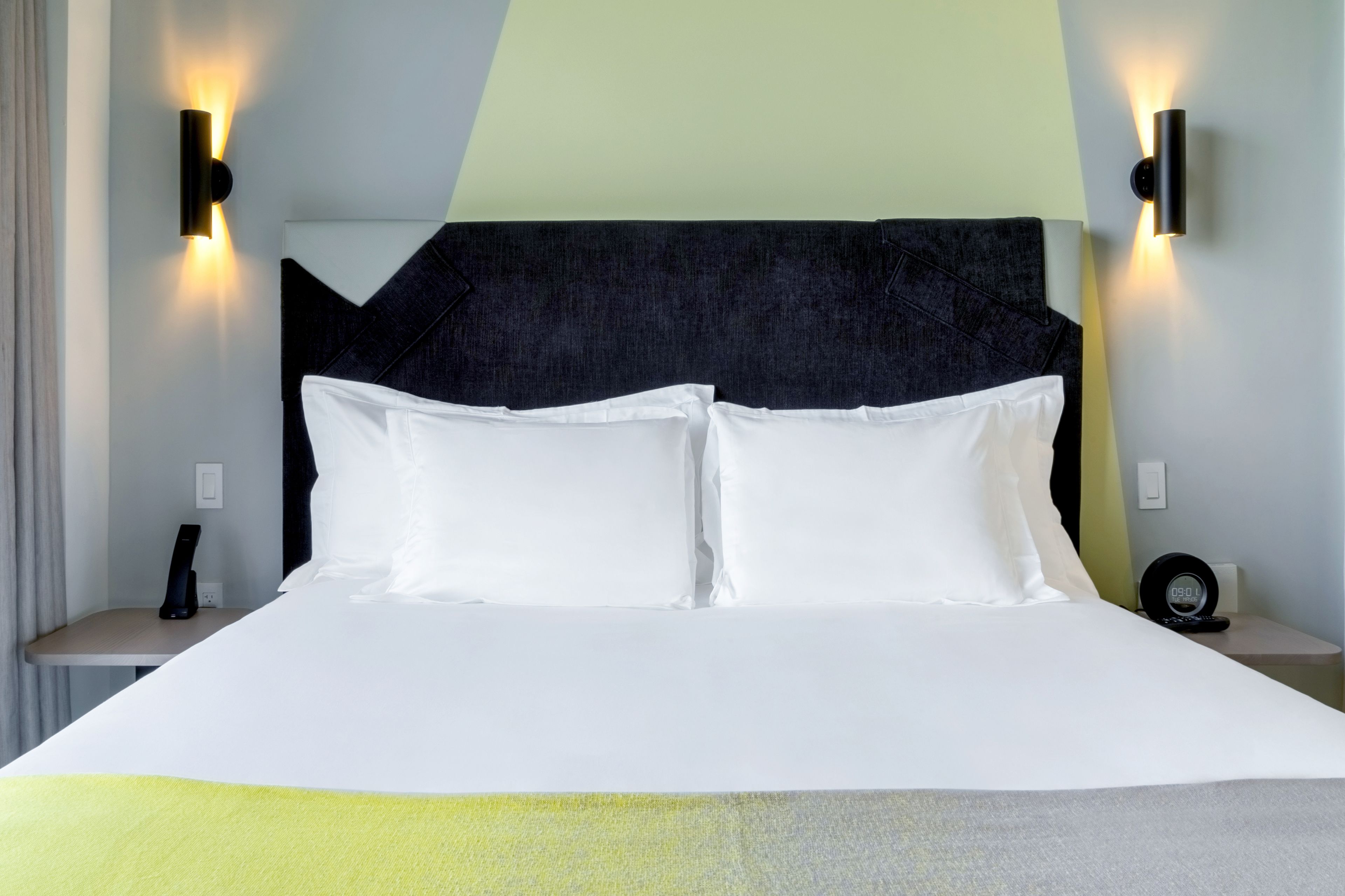 A neatly made bed with white linens, a dark headboard, and two wall-mounted nightstands with a clock and a phone on one side.