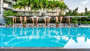 Ein Swimmingpool mit Liegestühlen und Sonnenschirmen, umgeben von Palmen und einem modernen Gebäude.