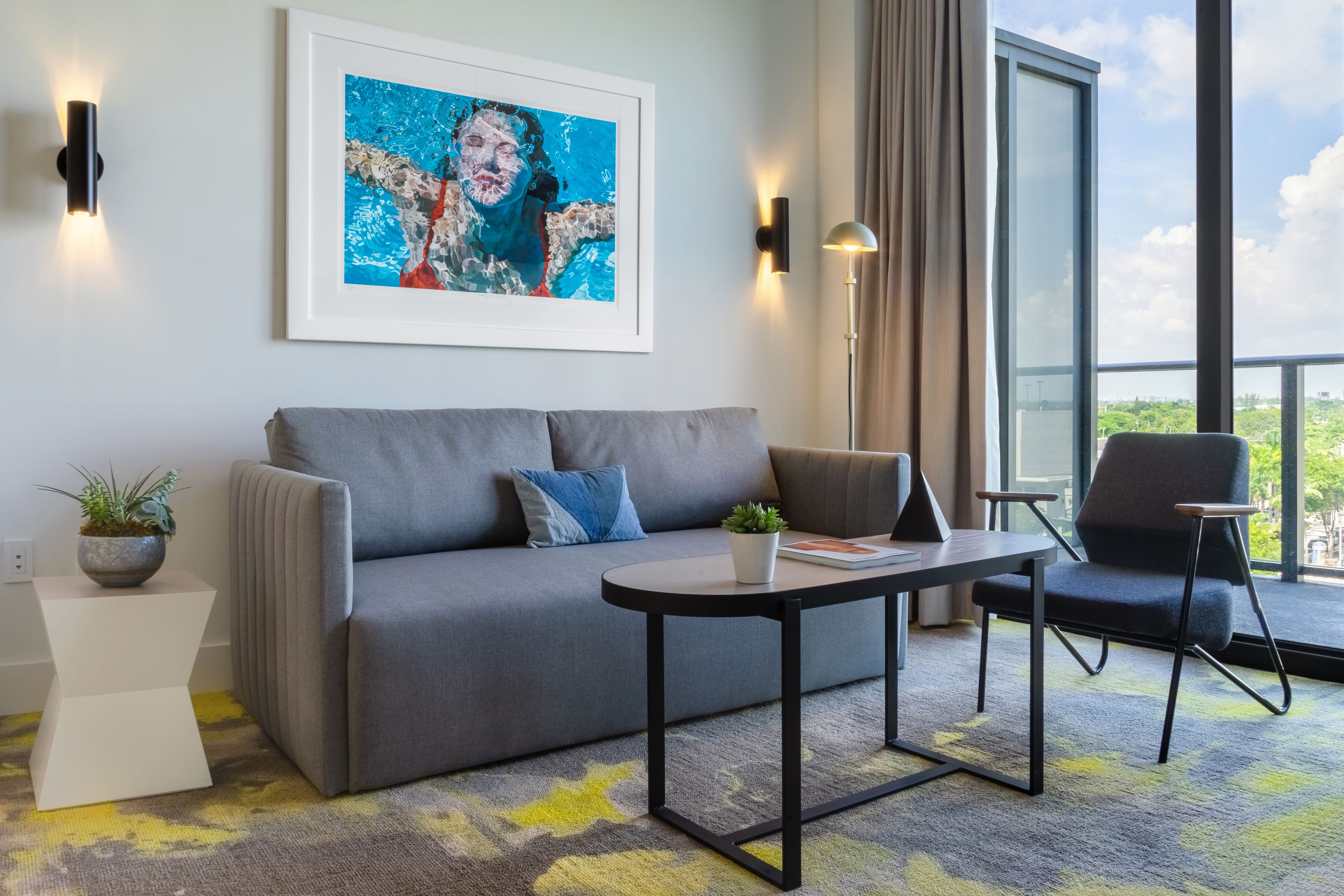 Et moderne hotelværelse med en grå sofa, et lille bord, en sort stol, et indrammet billede og et stort vindue med udsigt over byen.