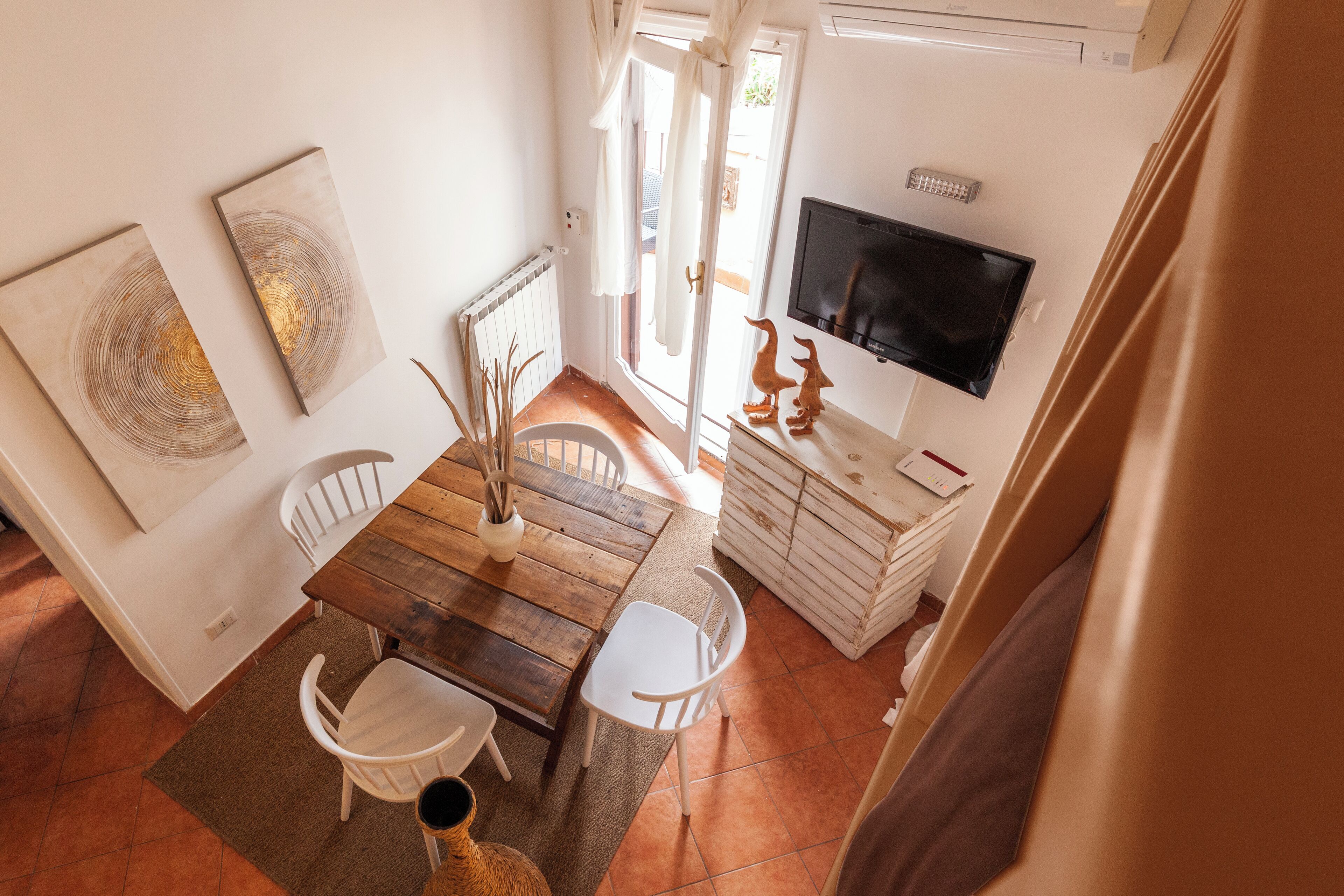 Un comedor con mesa de madera y sillas blancas, televisor montado en la pared y ventana con cortinas.
