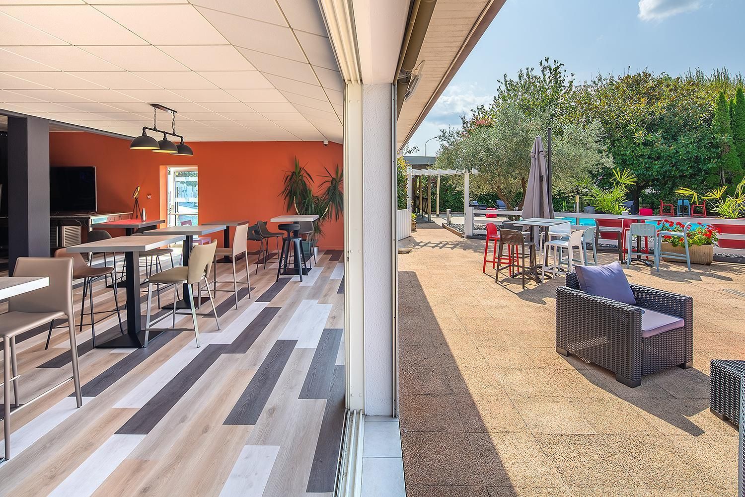 Un área de comedor al aire libre con varias mesas y sillas, un espacio de estar cubierto y vistas a un jardín con diversas plantas y una cerca.