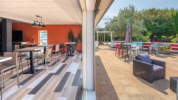 An outdoor dining area with multiple tables and chairs, a covered seating area, and a view of a garden with various plants and a fence.