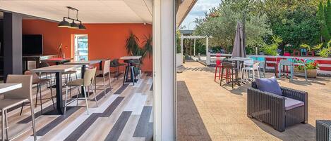 Un área de comedor al aire libre con varias mesas y sillas, un espacio de estar cubierto y vistas a un jardín con diversas plantas y una cerca.