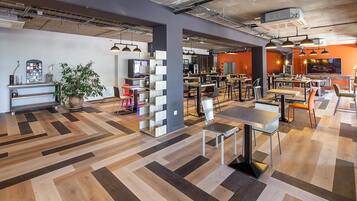A modern restaurant with a herringbone wood floor, exposed ceiling pipes, and a mix of seating arrangements including tables and chairs.