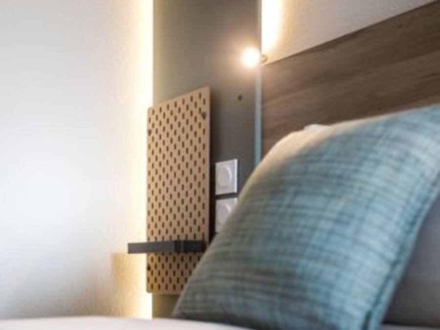 A modern hotel room with a wooden headboard, a bedside table with a lamp, and a patterned wall panel.