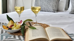 Una cama bien hecha con sábanas blancas, dos copas de vino blanco, un cuenco de fruta y un libro abierto sobre un mantel estampado.