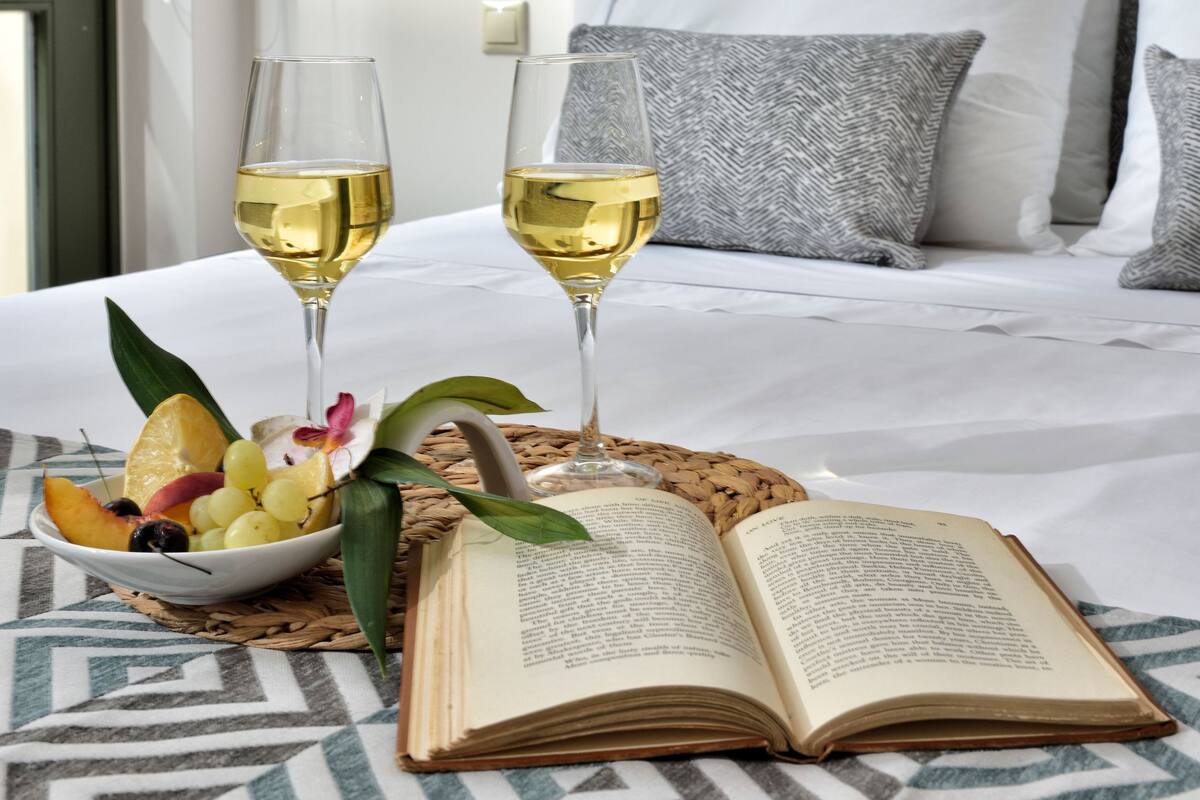 Un lit bien fait, avec du linge de lit blanc, deux verres de vin blanc, un bol de fruits et un livre ouvert sur une nappe à motifs.
