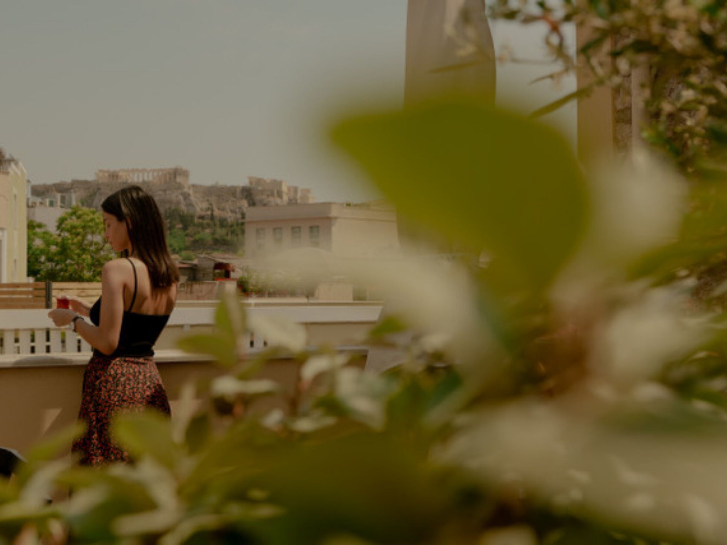 Une personne se tient sur un balcon, d’où elle a une vue sur un paysage urbain composé d’immeubles et d’espaces verts.