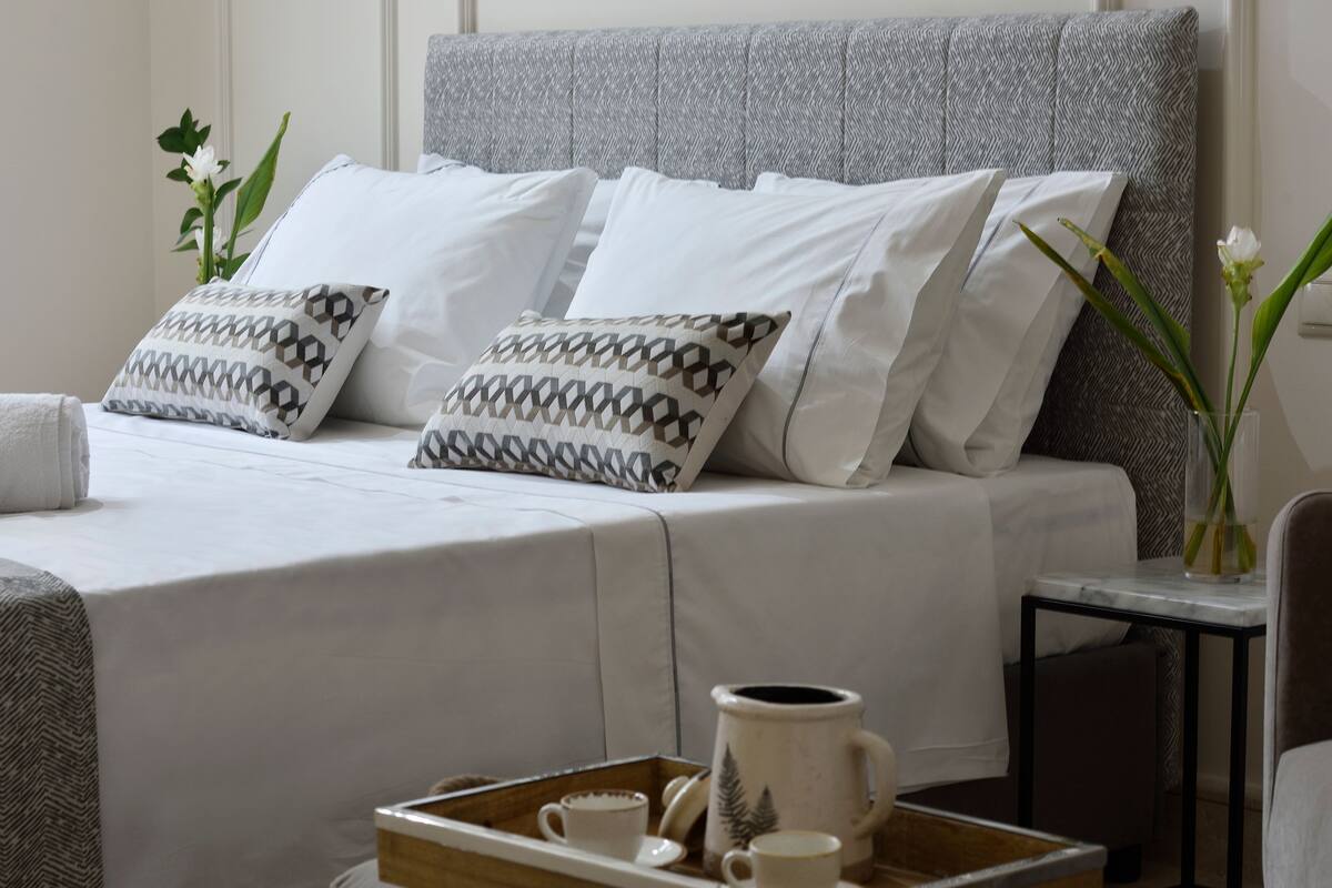 Un lit bien fait, avec du linge de lit blanc et des coussins à motifs, un plateau avec des tasses et une tasse, et un vase rempli de fleurs sur une table de chevet.