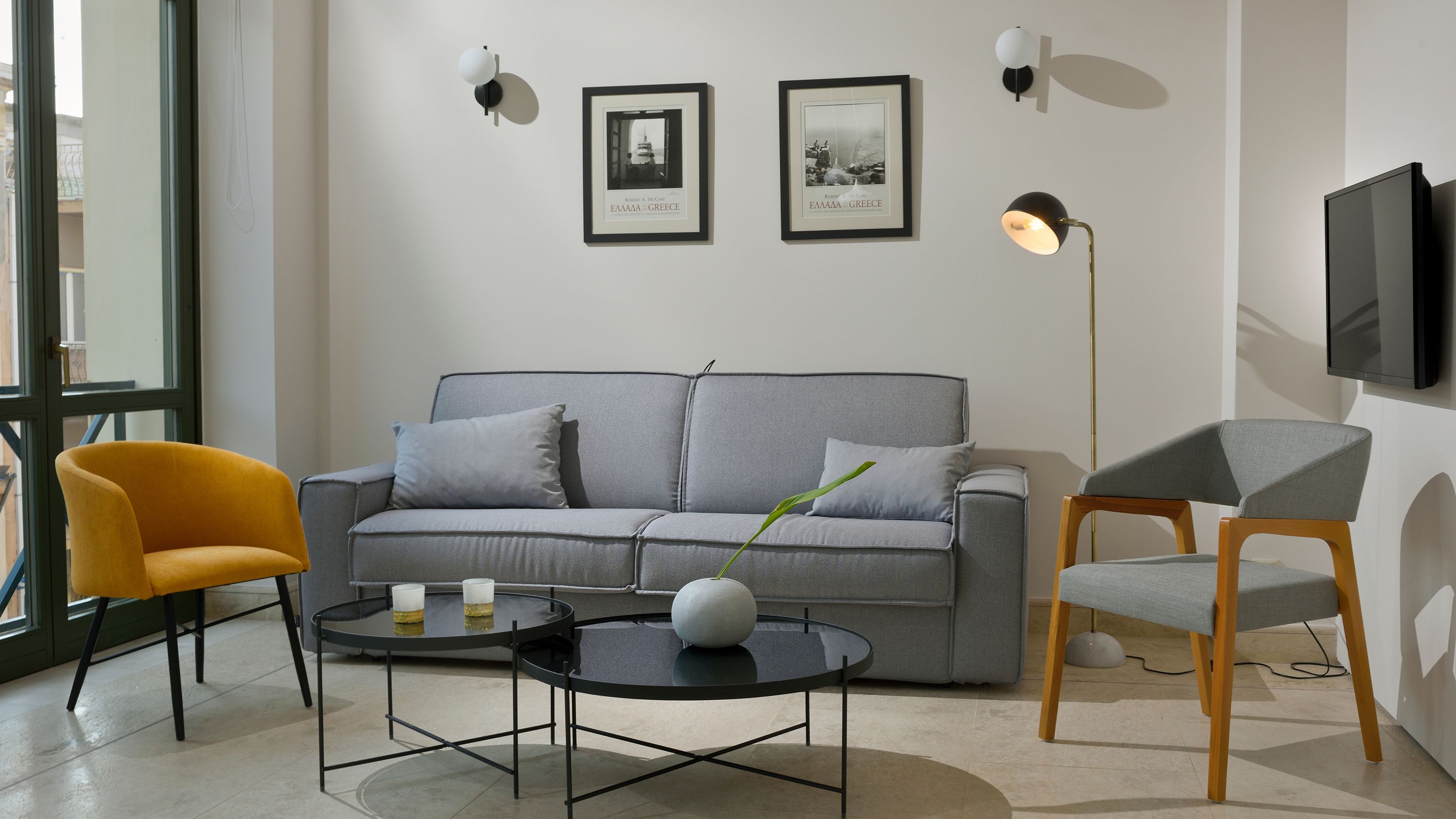 Ett modernt vardagsrum med en grå soffa, en gul fåtölj, ett runt soffbord och en golvlampa. På väggen hänger två inramade tavlor och en tv är monterad på väggen.