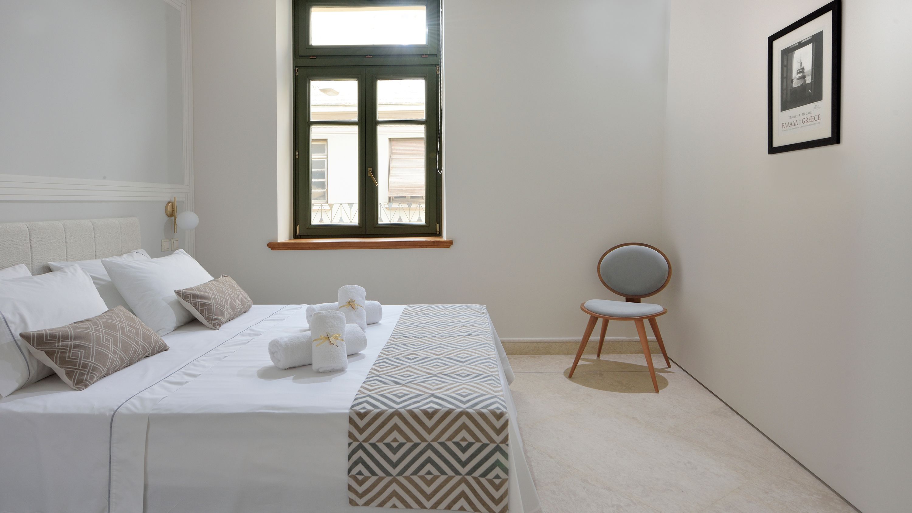 A neatly arranged bedroom with a bed, pillows, a chair, a window, and a framed picture on the wall.