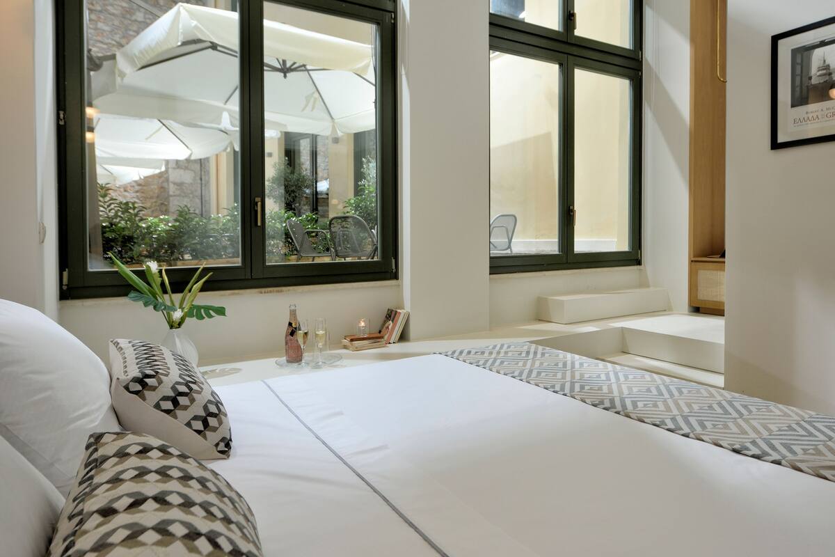 Une chambre d’hôtel avec un grand lit, une petite table sur laquelle se trouve un vase et deux verres, et une fenêtre donnant sur l’extérieur.