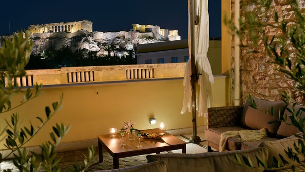 Un balcón con vistas a ruinas antiguas de noche, una mesa con velas y asientos al aire libre.