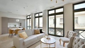 Una sala moderna con un sofá, sillones, una mesa de centro y un comedor con una mesa y sillas. Amplias ventanas ofrecen vistas a la calle.