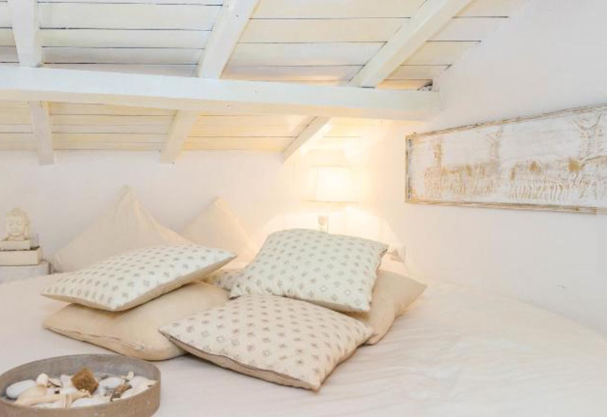 Un dormitorio con cama, almohadas, una mesita con un cuenco de conchas marinas y un cuadro en la pared.