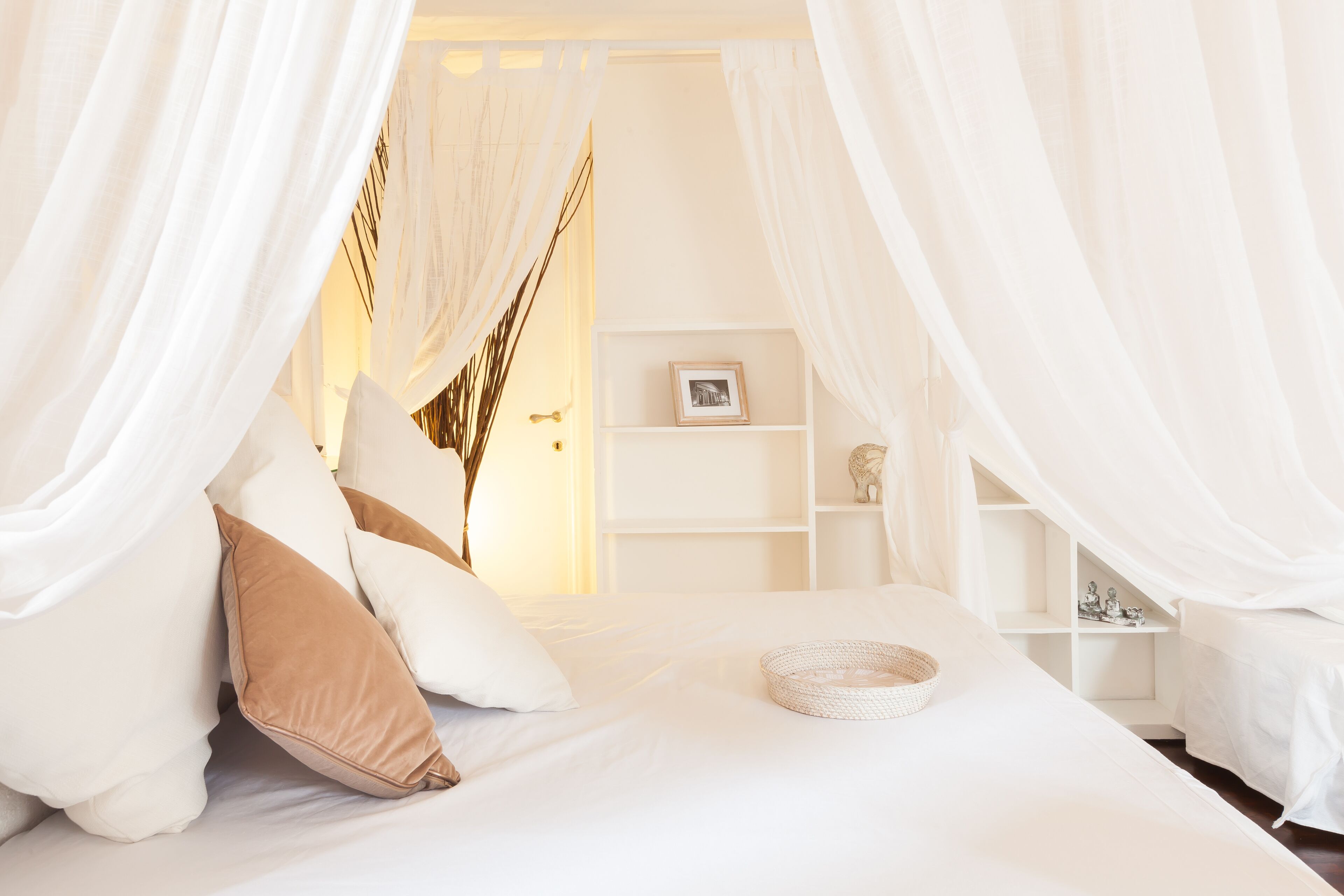 A bedroom with a canopy bed, white bedding, and a bedside table with a basket.