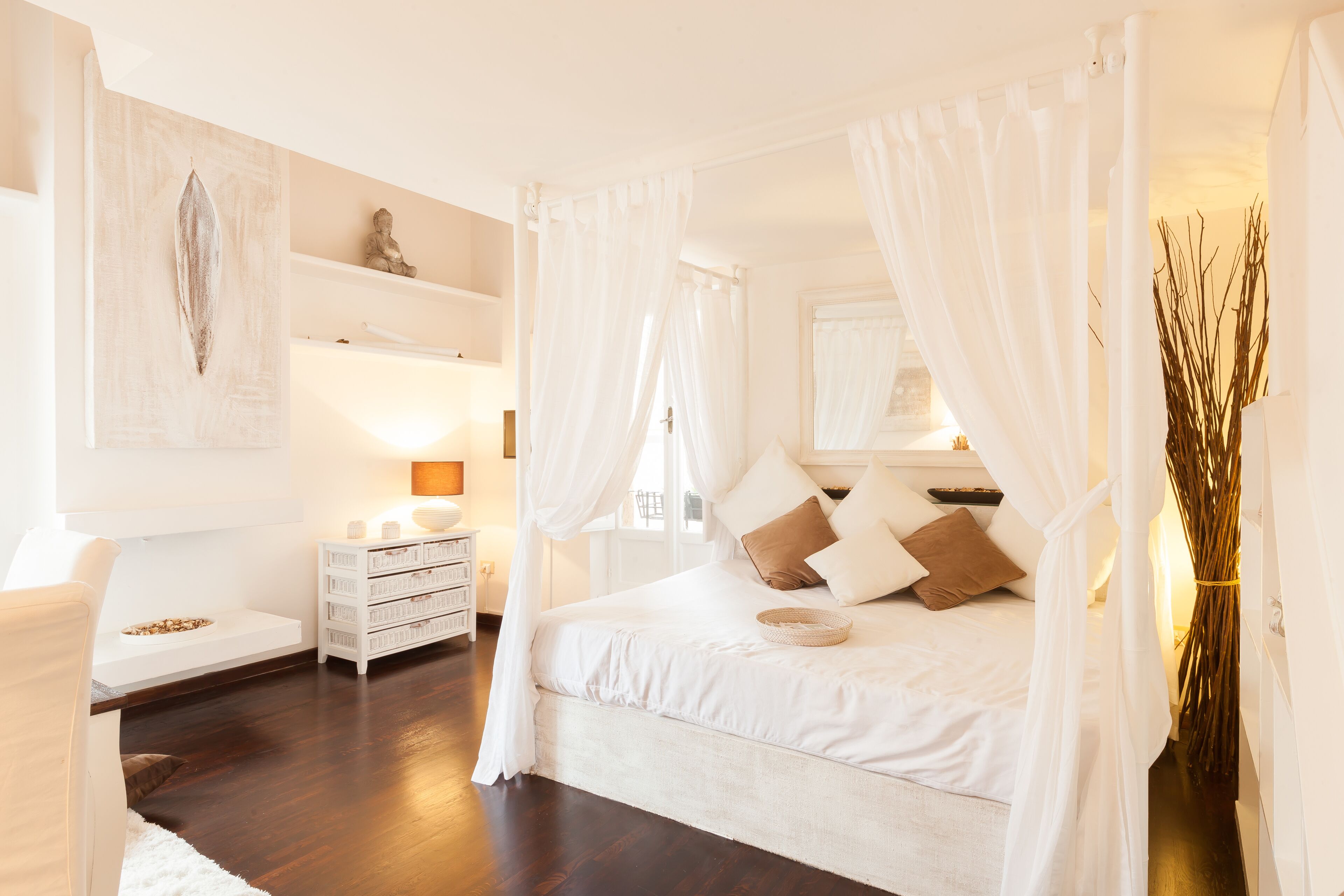 A bedroom with a large bed, white curtains, a bedside table with a lamp, and a wall-mounted shelf with decorative items.