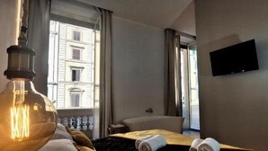 A hotel room with a large window, a flat-screen TV mounted on the wall, a bedside table, and a bed with a rolled-up towel. - Vatican Rome Suite (Rome)