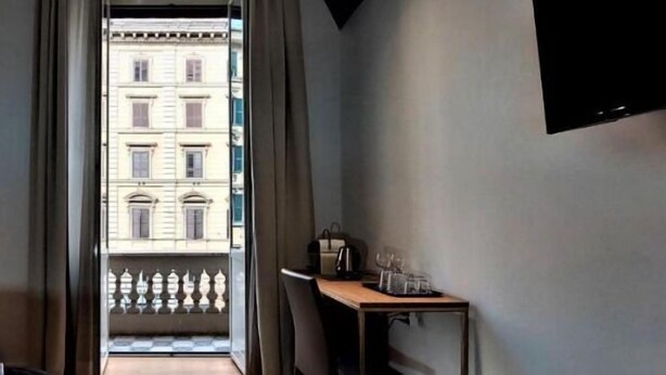 A room with a view of a building through a window, a chair, a small table with a kettle and glasses, and a wall-mounted TV. - Vatican Rome Suite (Rome)