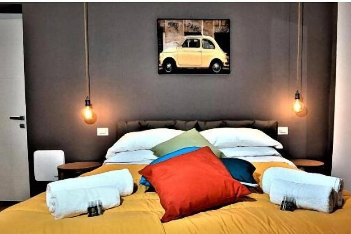 Camera d'albergo con un letto, cuscini, un comodino, un quadro appeso al muro e una porta.