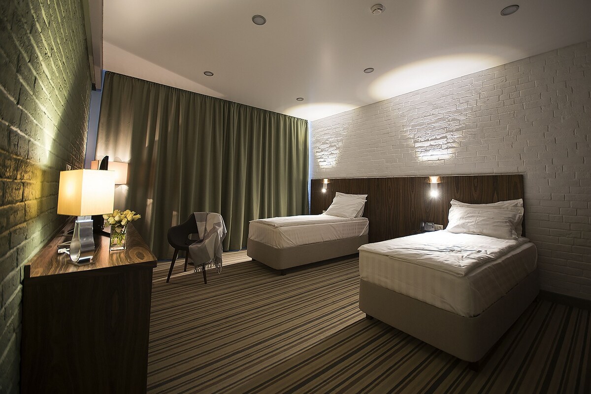 Habitación de hotel con dos camas, un escritorio con lámpara, una silla y una pared de ladrillo.