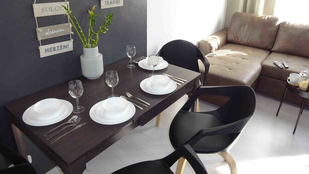 Ein Essbereich mit einem dunklen Holztisch, gedeckten Plätzen für vier Personen, weißen Tellern und Weingläsern. Zwei hängende Schilder mit deutschem Text, eine weiße Vase mit Grünpflanzen und ein beiges Sofa im Hintergrund.