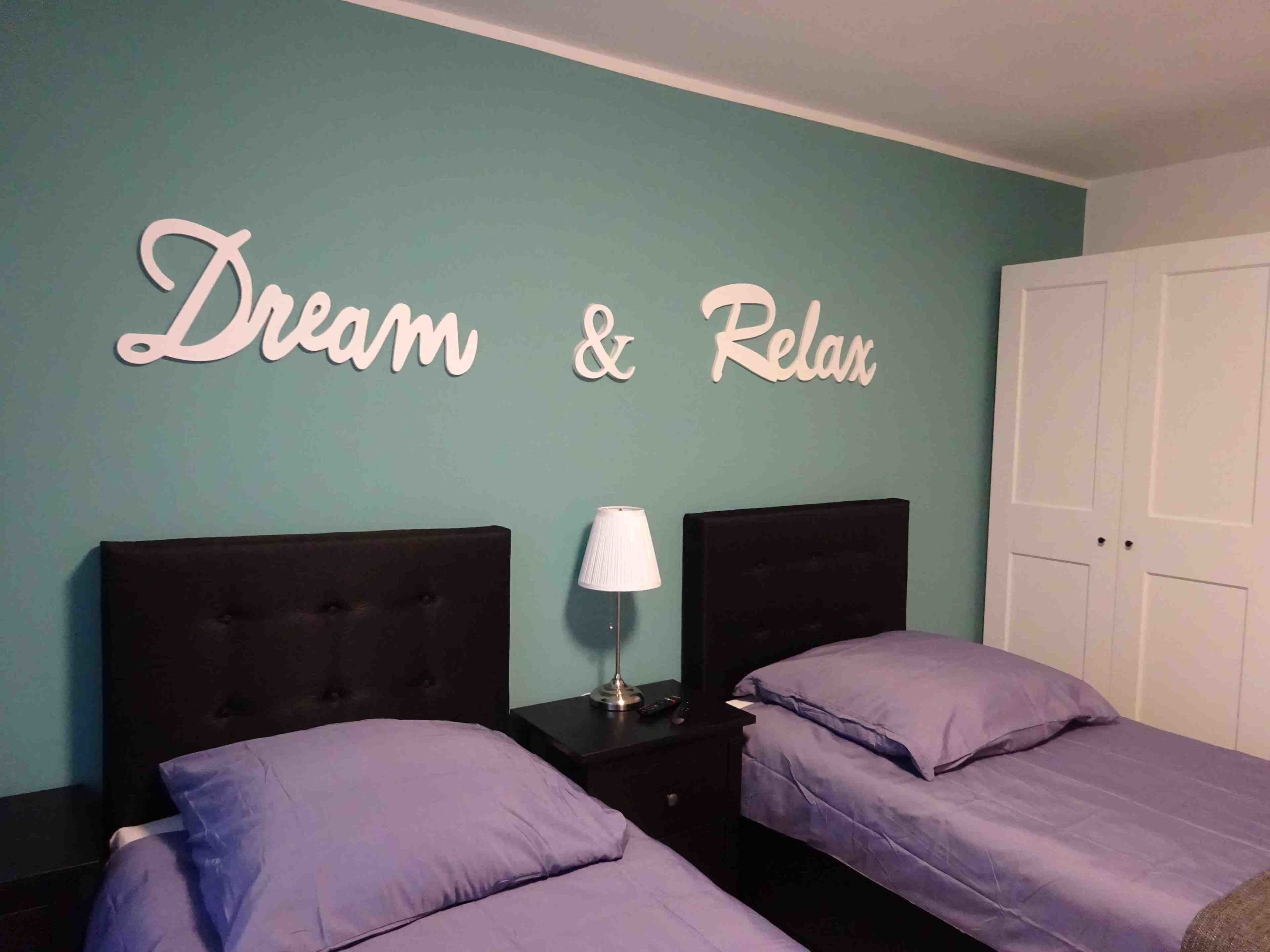 A bedroom with two beds, a nightstand with a lamp, and a wall sign that reads "Dream & Relax".