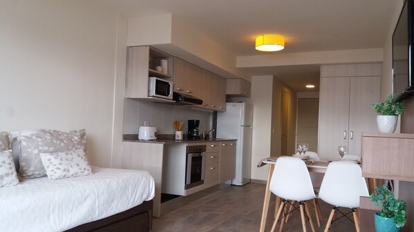 A compact bedroom with a bed, a small kitchenette, and a dining area with chairs and a table.