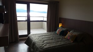 A hotel room with a bed, a nightstand with a lamp, and a view of a body of water through a large window.