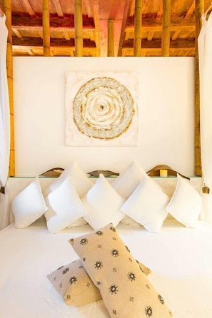 Una cama con almohadas blancas y estampadas, techo de madera y una obra de arte circular en la pared.