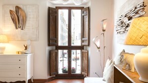 Una habitación con un ventanal grande, un tocador blanco, una lámpara y una estantería con adornos.