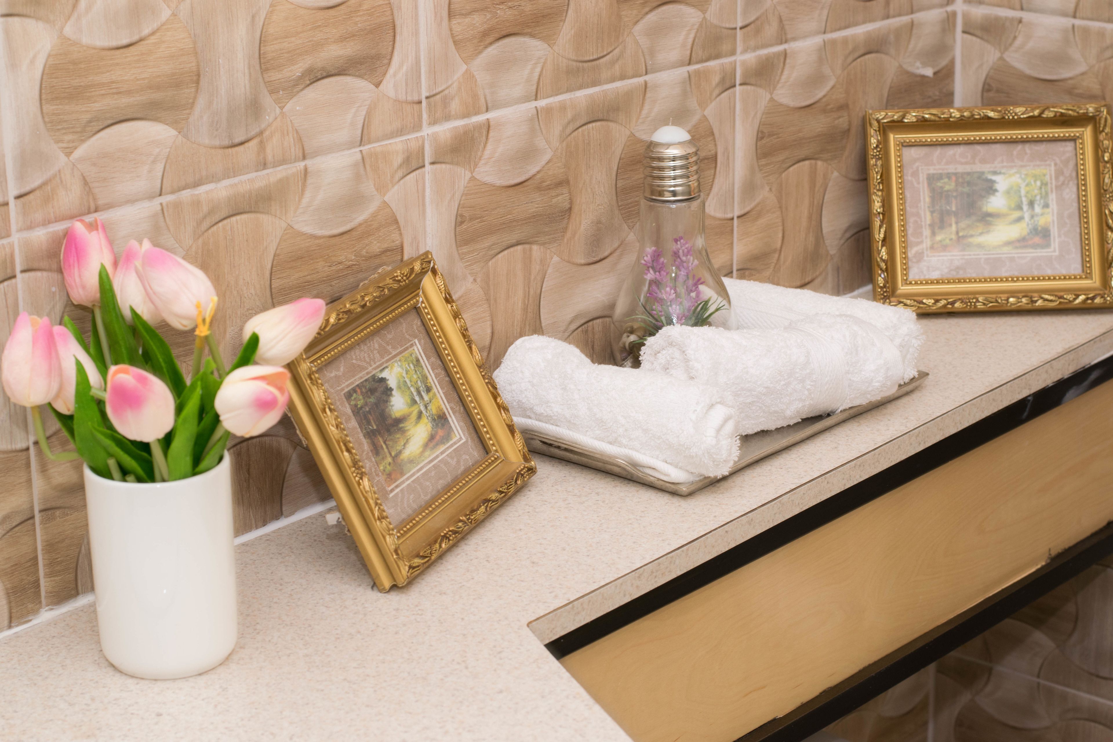 Un meuble-lavabo de salle de bain agrémenté d’un vase blanc contenant des tulipes roses, d’un cadre photo doré, d’une bouteille en verre avec un bouchon blanc et d’une serviette blanche pliée.