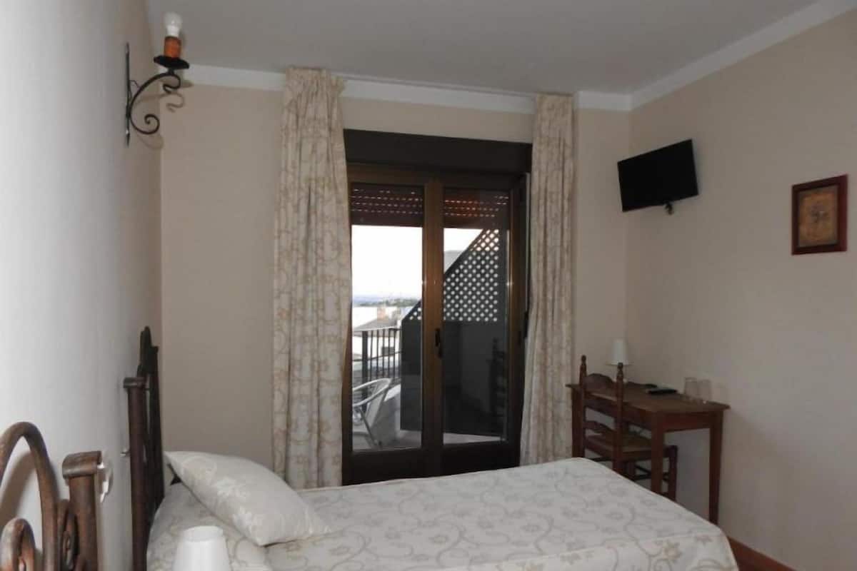 A bedroom with a bed, a desk, a chair, a TV, and a balcony with a view.