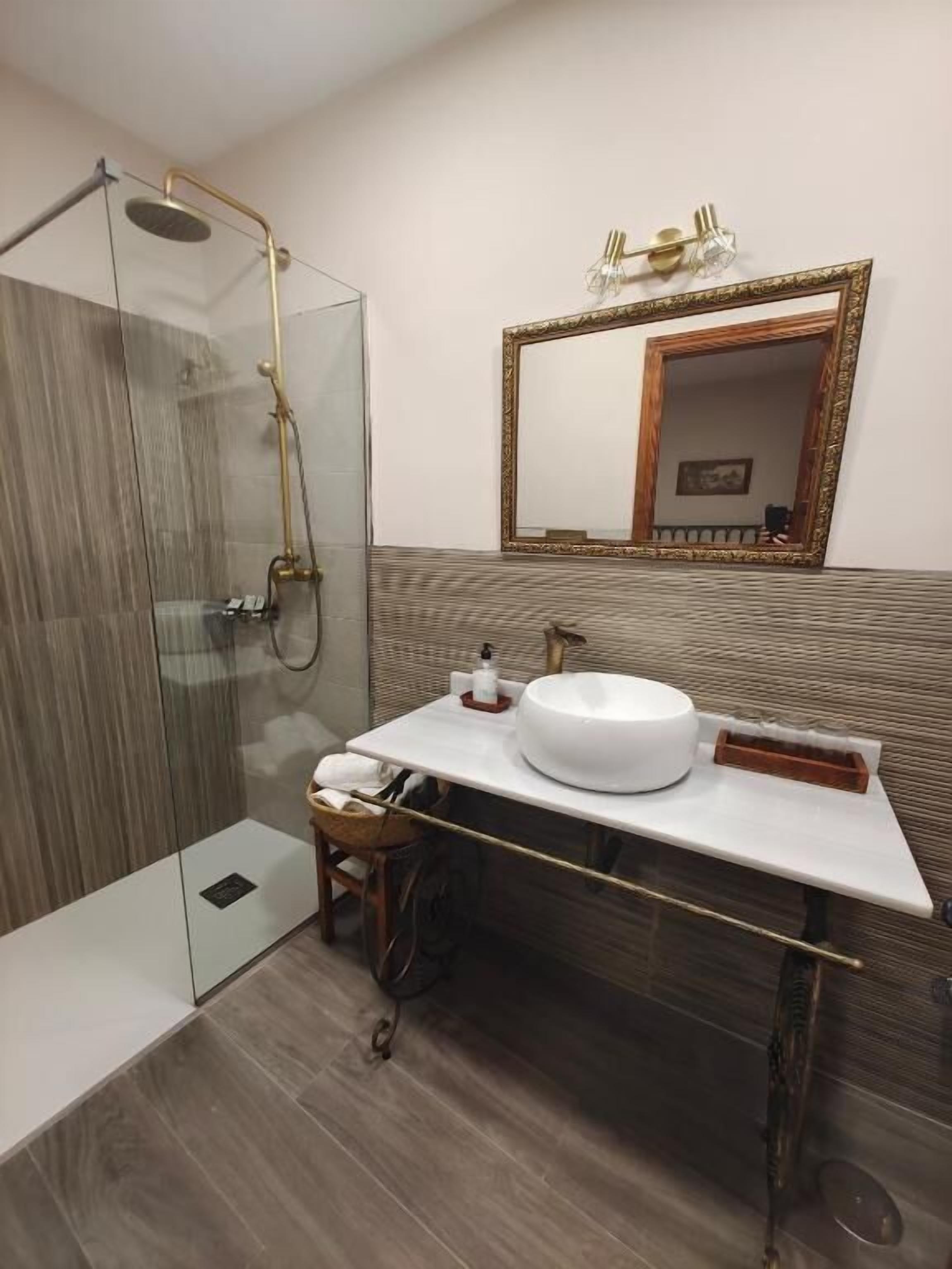 Une salle de bain moderne avec une douche à parois de verre, un lavabo blanc doté d’un bac rond et un grand miroir avec un cadre doré.