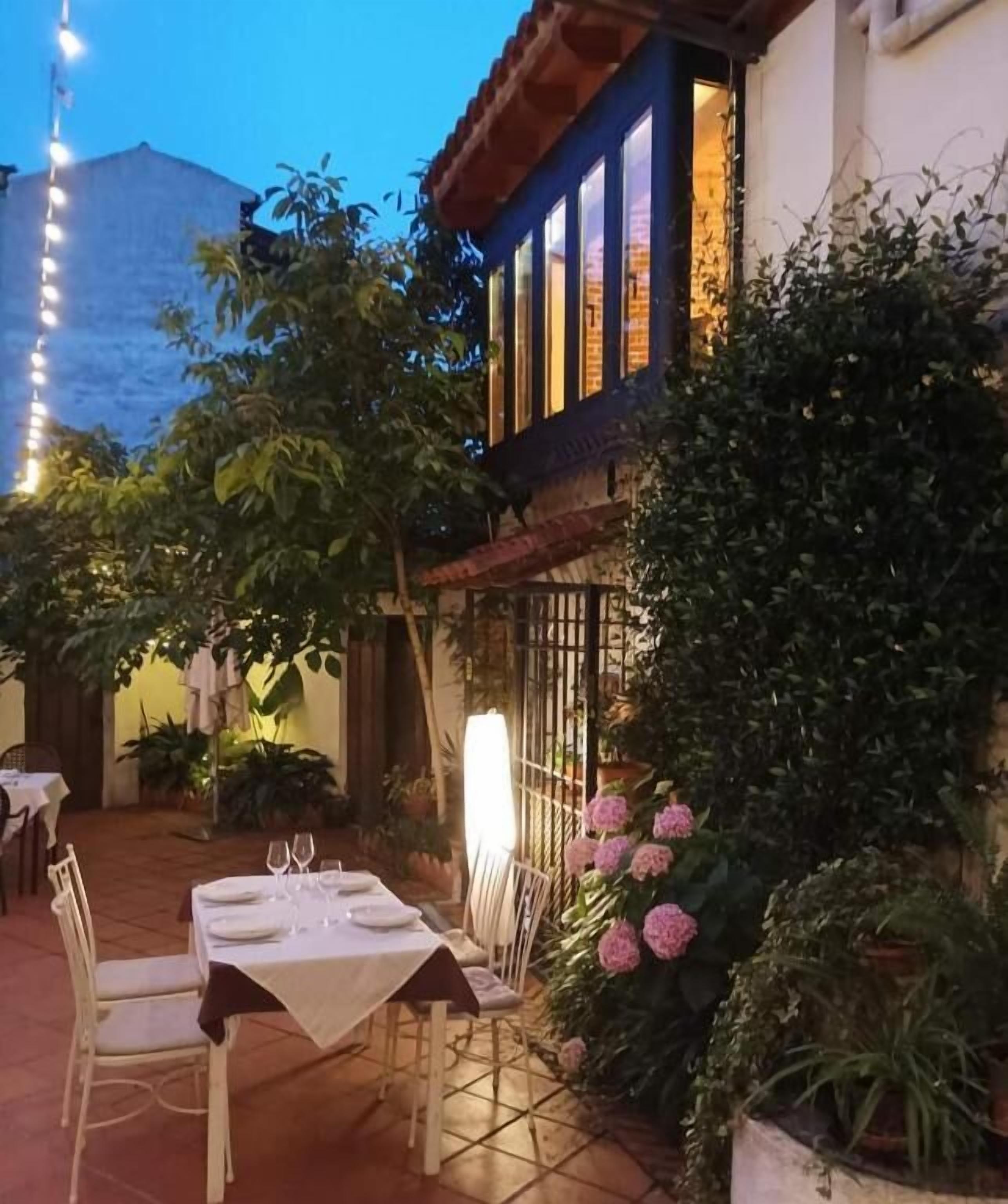 An outdoor dining area with tables set for two, surrounded by potted plants and flowers.