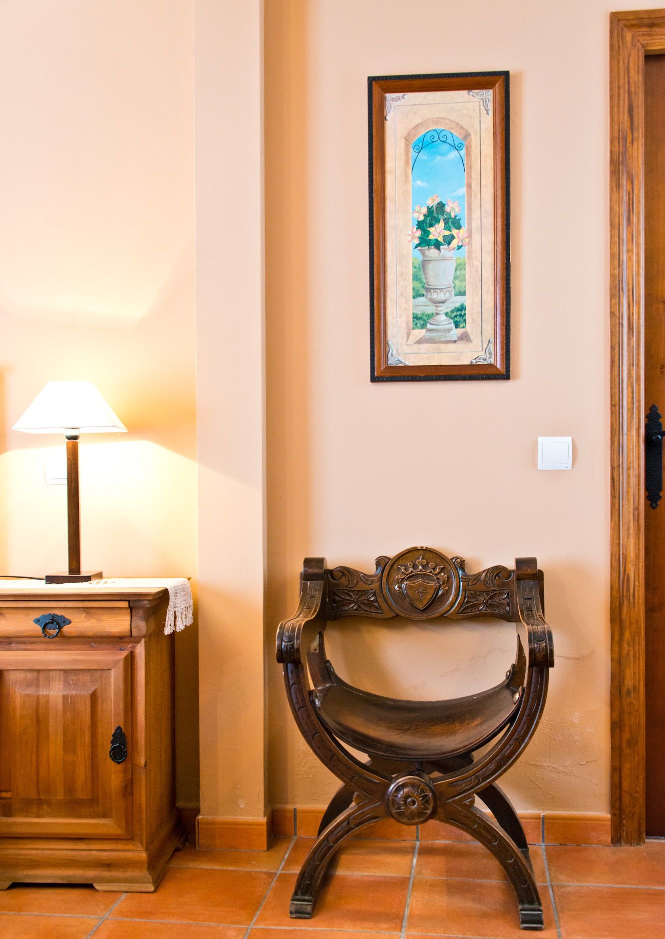 Un banco de madera con detalles de metal labrado, un cuadro enmarcado encima y una lámpara sobre una mesa de madera a la izquierda.