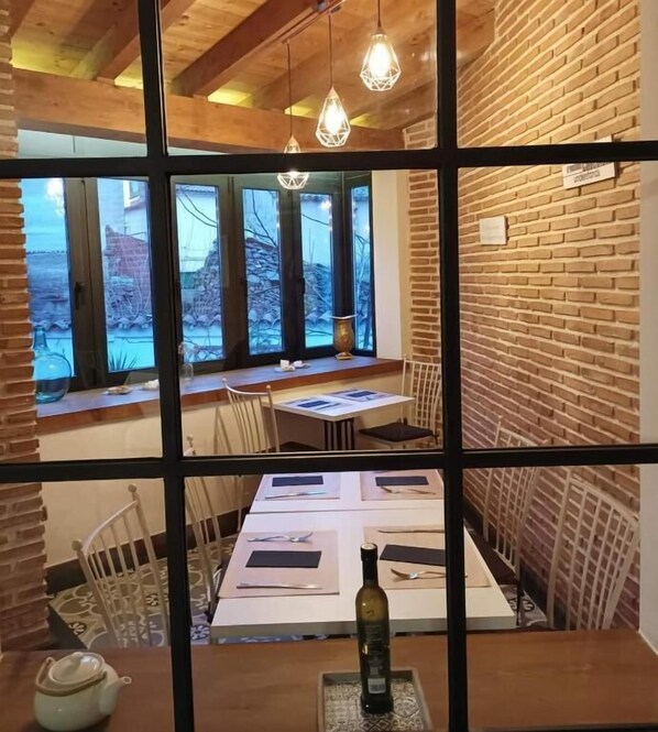 Un comedor con pared de ladrillo, techo de madera y lámparas colgantes. Las mesas están preparadas con manteles individuales y cubiertos.