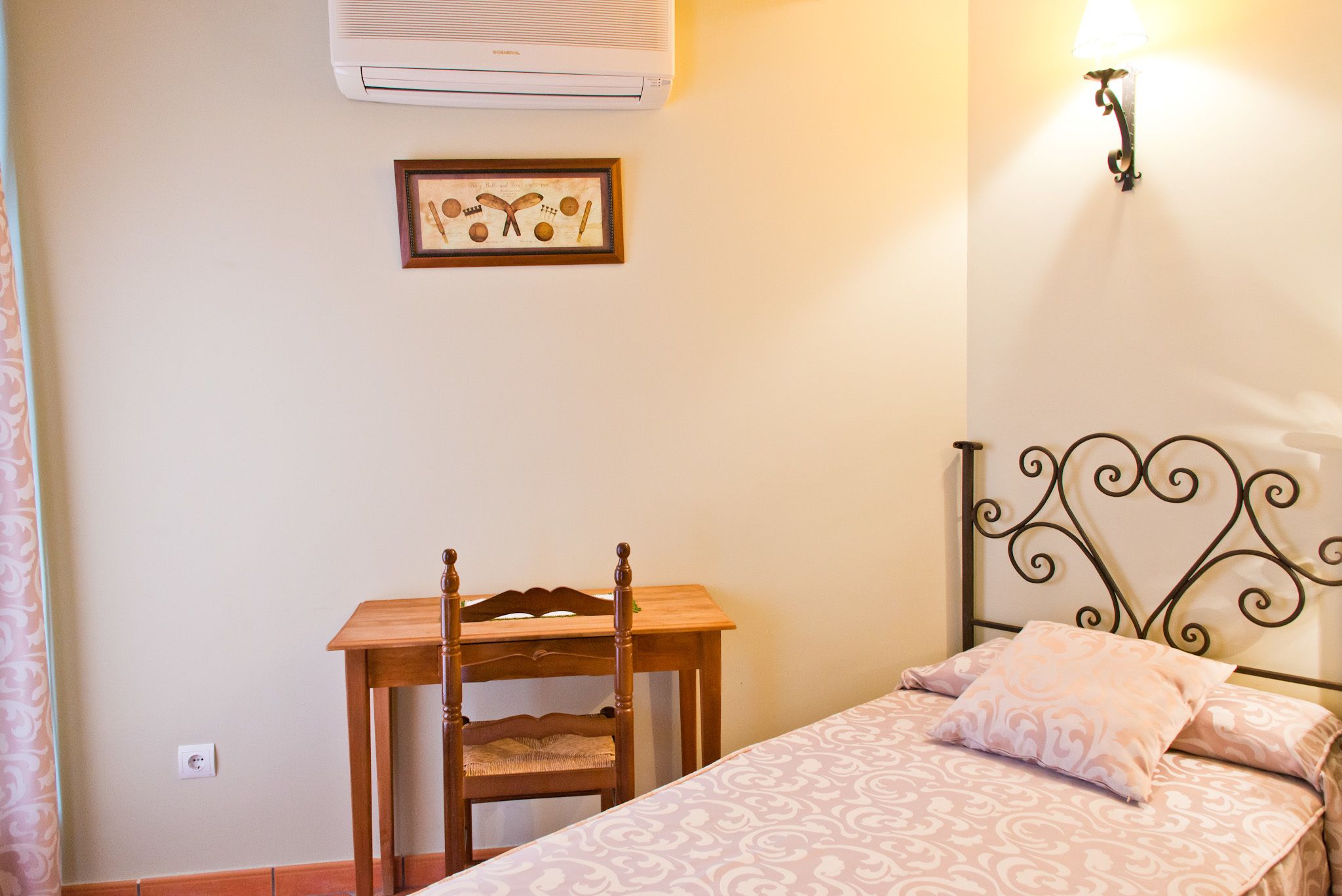 A bedroom with a bed, a wooden chair, a small table, a wall-mounted air conditioner, and a framed picture.
