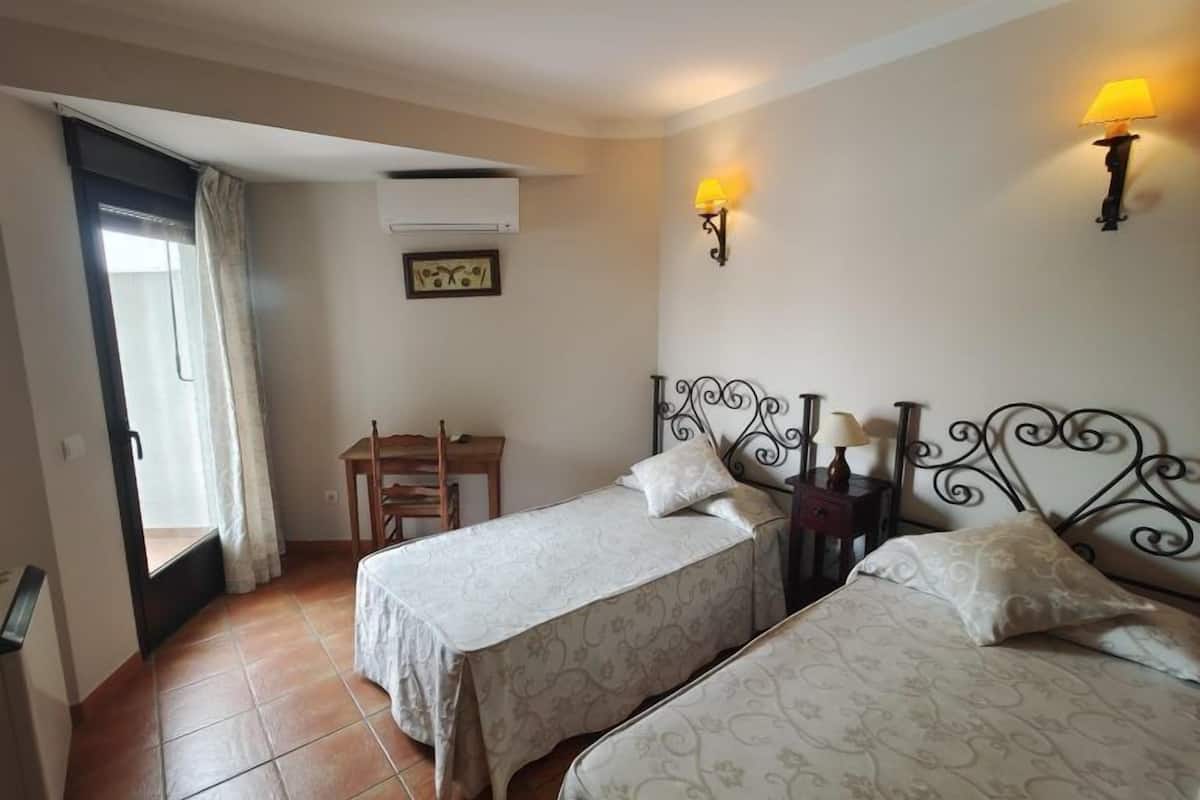 A room with two beds, a desk, a chair, a window with curtains, an air conditioning unit, and wall-mounted lamps.