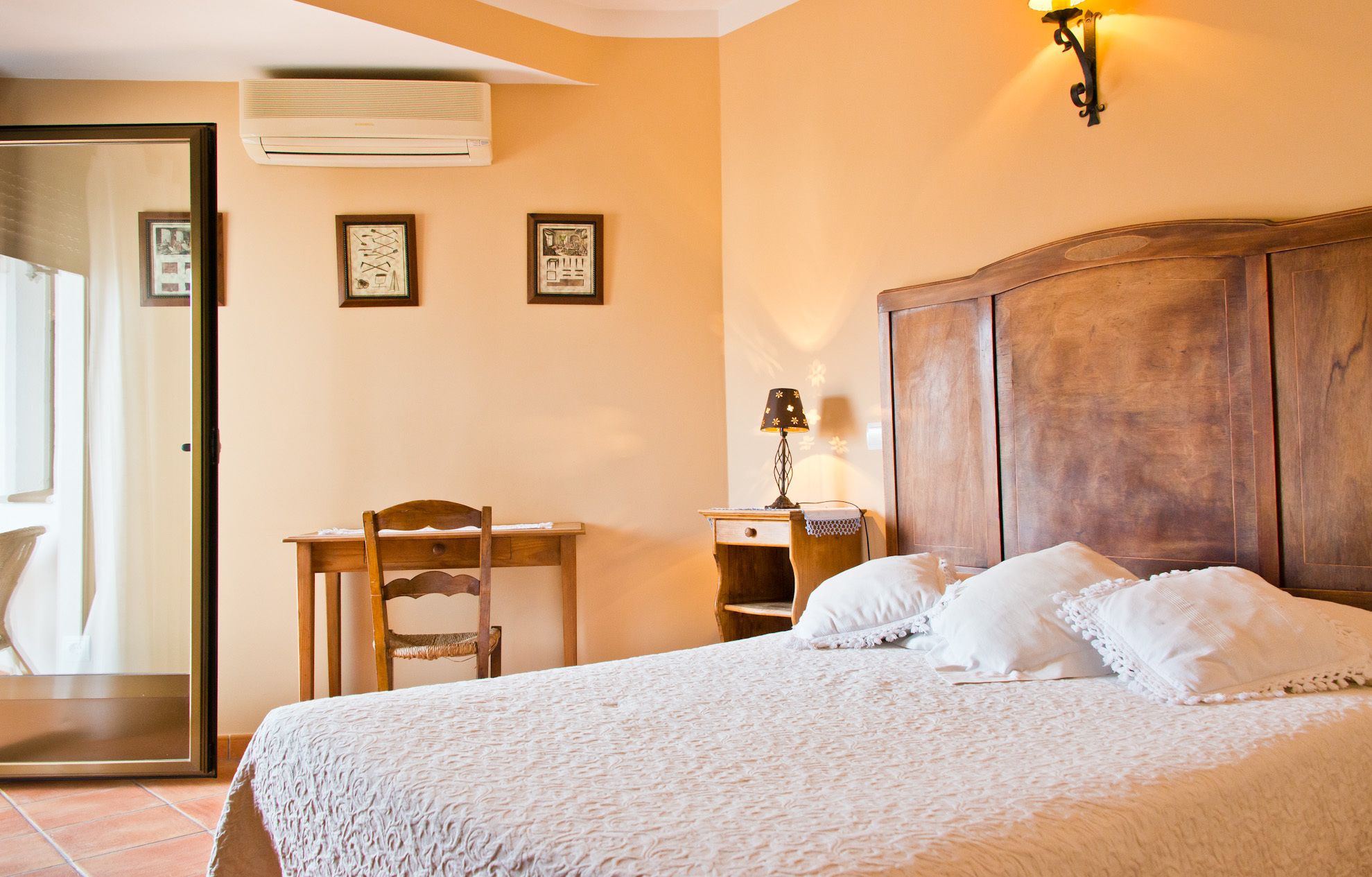 A bedroom with a bed, a desk, a chair, a nightstand, a lamp, and a wall-mounted air conditioner.