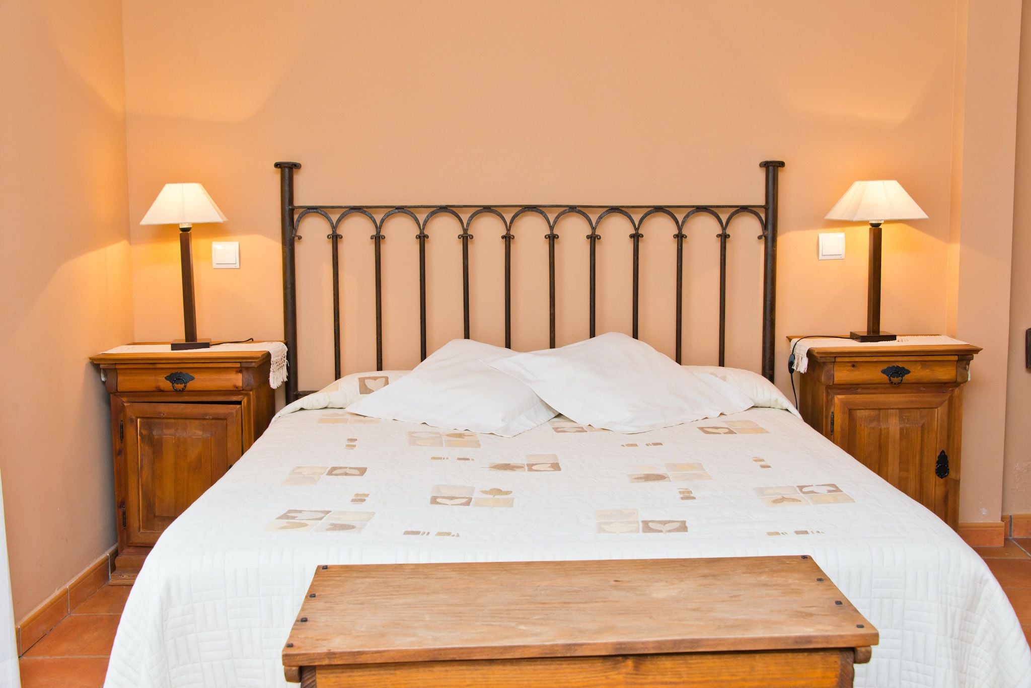 A neatly made bed with a patterned bedspread, two bedside tables with lamps, and a wooden bench in front of the bed.