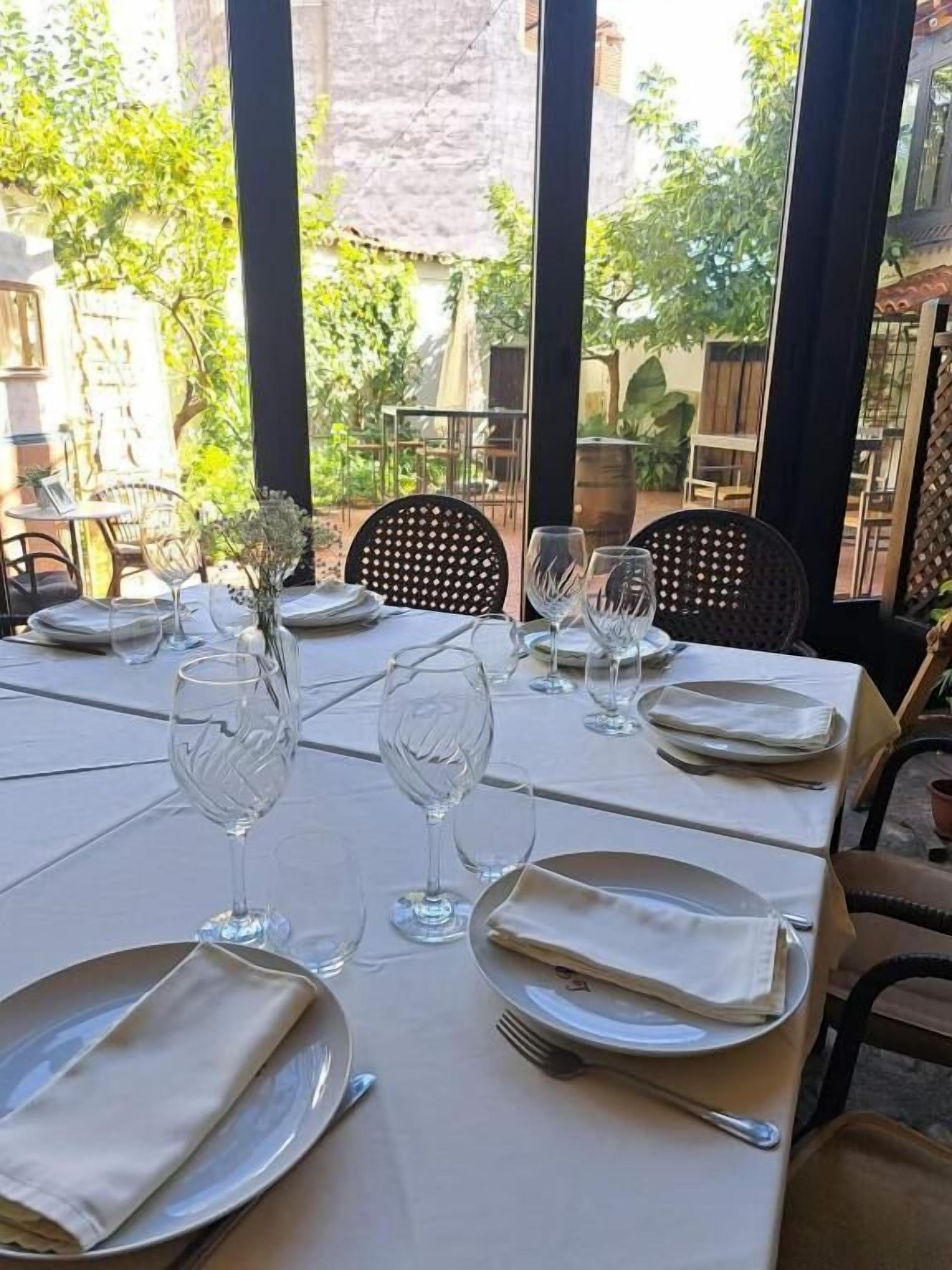 Mesa lista para comer, con platos, vasos y cubiertos, con vistas a un jardín.