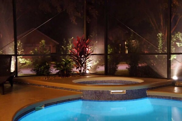 A swimming pool with a tiled edge, surrounded by plants and a view of a house through large windows.