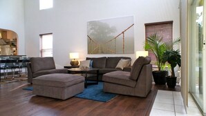 A spacious living room with a large window, a sofa set, a coffee table, and a painting of a bridge. - Shepherds Delight Apartment 5 Bedroom (Haines City)