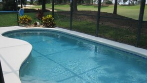 A swimming pool with a curved edge, surrounded by a metal fence and a grassy area with trees.