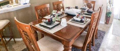 A dining area with a wooden table and chairs, a kitchen in the background, and a potted plant.