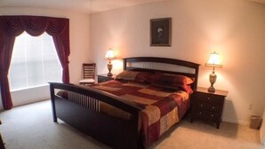 Ein Schlafzimmer mit einem Bett, zwei Nachttischen mit Lampen, einem Sessel, einem Fenster mit Vorhängen und einem gerahmten Bild an der Wand.